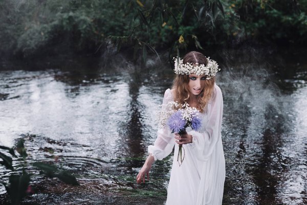 A beautiful girl in a white dress is walking along the river. In her hands she holds a bouquet of flowers. On her head is a wreath of white flowers.