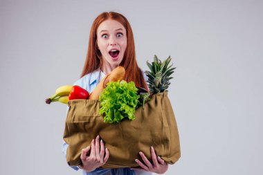 Muz, ananas, sebze gibi ürünlerle dolu bir çanta tutan kızıl saçlı güzel bir kadın.