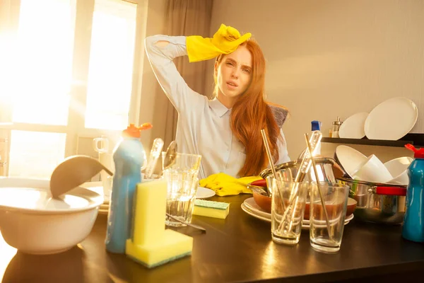 Δυστυχισμένη κοκκινομάλλα τζίντζερ όμορφη γυναίκα φορώντας κίτρινο γάντι και πλύσιμο σωρό βουνά από βρώμικα πιάτα: κύπελλα, τηγάνι, κατσαρόλα στην κουζίνα — Φωτογραφία Αρχείου
