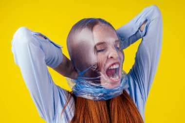 redhaired ginger european woman suffocation head in blue polyethylene package in studio yellow background. eco pollution cellophane film concept clipart