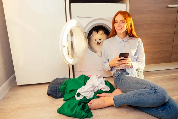 Eccitata divertente ragazza rossa ascoltare musica con le cuffie, il suo adorabile soffice spitz all'interno della lavatrice a casa un mucchio di vestiti sporchi sul pavimento — Foto Stock