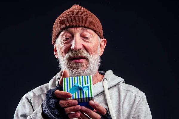 Kahverengi şapkalı ve sızdıran eldivenli yaşlı bir serserinin gülümseyen portresi, stüdyo siyah arka planda mavi bir hediye kutusu tutuyor.