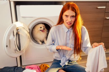 Fie, what stench Shocked excited redhaired girl wash pile of dirty smelly clothes ,her adorable fluffy spitz helpinh inside the washing machine at home
