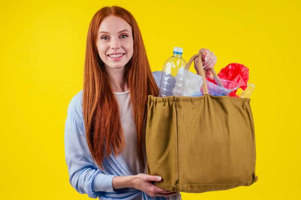 Красивая рыжеволосая счастливая женщина с мешком хлопка эко с сортировкой мусора: пластиковая бутылка, одноразовый пакет в студии желтый фон — стоковое фото