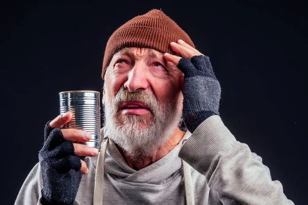 Retrato de triste anciano viejo vagabundo con sombrero marrón cálido y guantes con fugas, sosteniendo metal enlatado banco tarro para limosna de dinero en el estudio de fondo negro —  Fotos de Stock
