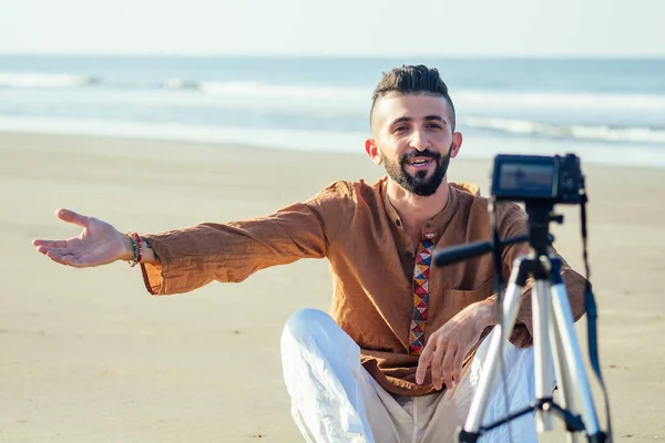 Арабский мужчина сидит на песке и говорит штатив видео решений на пляже. Концепция создания контента для путешествий — стоковое фото
