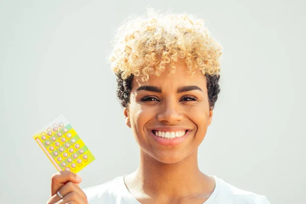 Fille brésilienne avec de jolies boucles afro blonde montrant des pilules contraceptives en studio fond blanc — Photo