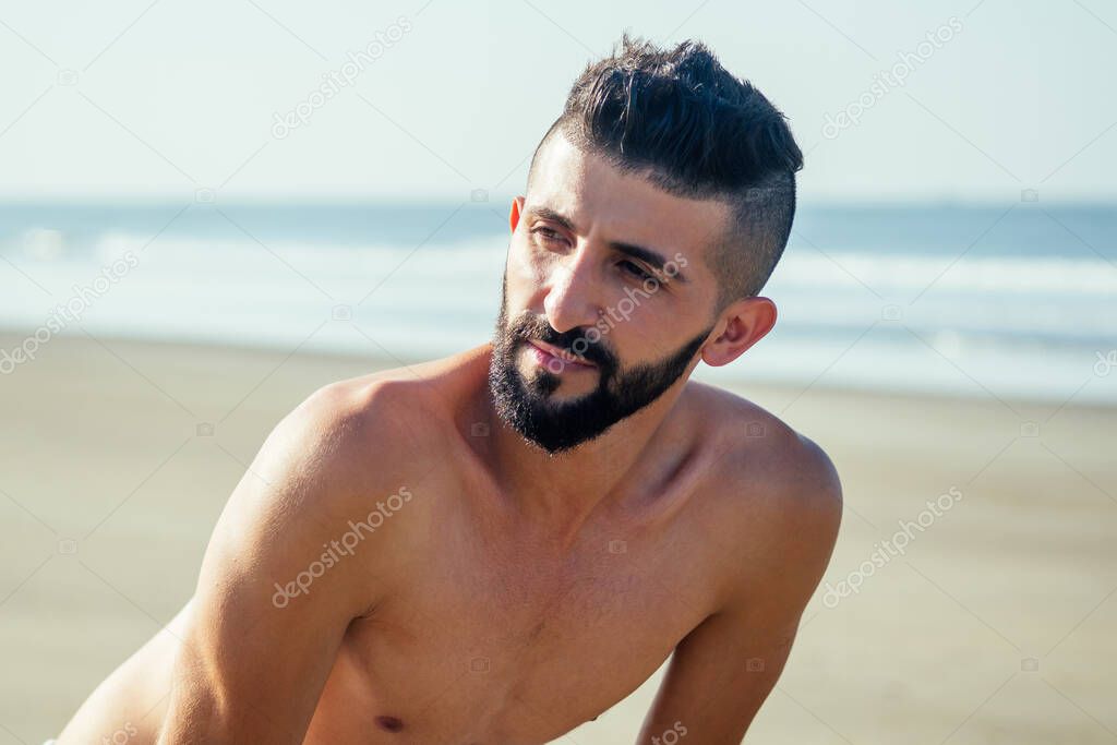 Young handsome indian man taking a rest after does push-ups on the beach in the morning. He tired and sweaty shirtless