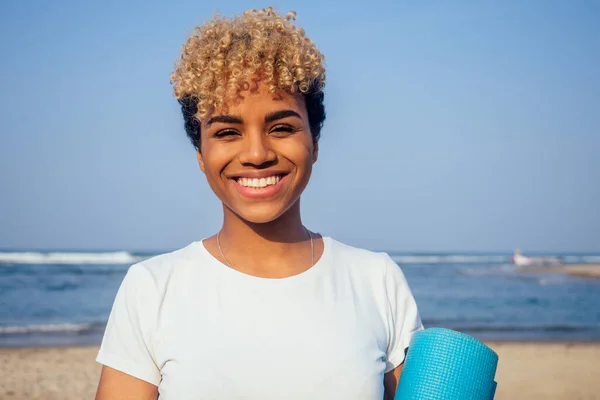 Jeune femme brésilienne portant un t-shirt blanc en coton décontracté et tenant un tapis de yoga sur la plage — Photo