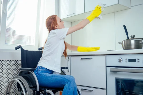 Genç, kızıl saçlı, tekerlekli sandalyede sakat bir kadın. Sarı kurdele eldiveni takıyor ve evi temizliyor. — Stok fotoğraf