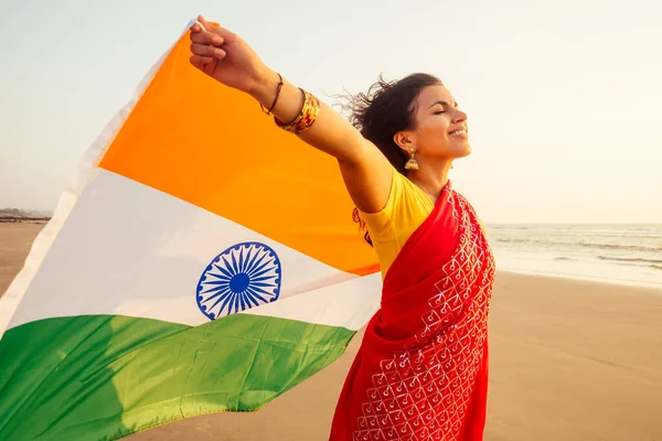 Bela mulher segurando bandeira indiana tricolor, vestindo sari tradição vermelha na praia do mar em Goa — Fotografia de Stock