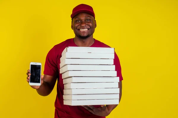 Afro-americký doručovatel s balíčkem v červeném tričku a čepici zobrazující obrazovku smartphonu ve studiu žlutém pozadí. Koncept aplikace Fast delivery food — Stock fotografie