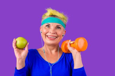 Şık sarı boyalı saçları olan neşeli, yaşlı bir kadın. Alnında mavi bir bandaj olan, turuncu dambılı olan ve spor salonunun arka planında kulaklıkla müzik dinleyen bir kadın.