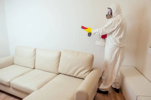 Trabajador de control de plagas en la pulverización uniforme de pesticidas debajo del sofá en la sala de estar — Foto de Stock