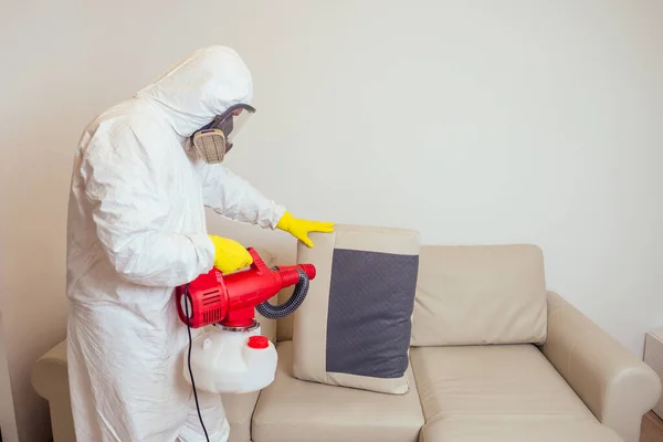 Lucrător de control al dăunătorilor în pesticide uniforme de pulverizare sub canapea în camera de zi — Fotografie, imagine de stoc