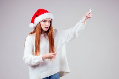 Kızıl saçlı kızıl saçlı kadının kırmızı Noel Baba şapkası ve örgü süveter giymesine şaşırmış ve şaşırmış halde stüdyo beyaz arka planda uzun bir alışveriş makbuzu tutuyor. büyük para xmas harcamaları