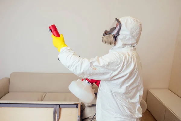 Εργάτης καταπολέμησης παρασίτων με στολή ψεκασμού φυτοφαρμάκων κάτω από καναπέ στο σαλόνι — Φωτογραφία Αρχείου