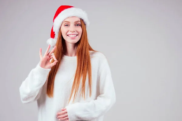 Schöne rothaarige Ingwerfrau mit weißem Strickpullover und Weihnachtsmann-Mütze im Studio-Hintergrund — Stockfoto