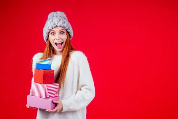 Rothaarige Ingwerfrau trägt stylische Wollmütze und weißen Pullover, blickt in die Kamera und hält drei Schachteln mit Geschenken in Studio-rotem Hintergrund — Stockfoto