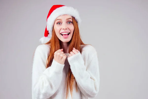 Vacker rödhårig ingefära kvinna bär vit stickad tröja och Santa claus hatt finger kors gör önskningar i studio bakgrund — Stockfoto