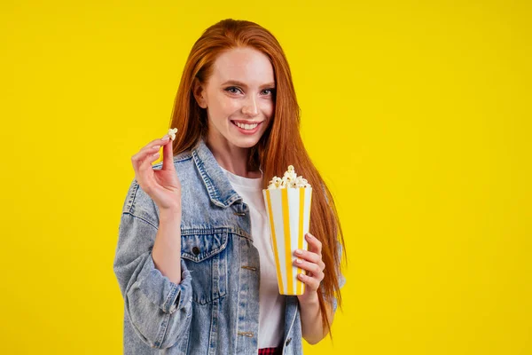 Emotionale rothaarige Ingwerfrau trägt Jeansjacke und hält Popcorn im Studio gelben Hintergrund — Stockfoto
