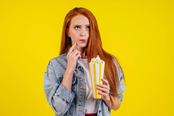 Emotionale nachdenkliche Kalorien rothaarige Ingwerfrau trägt Jeansjacke und hält Popcorn im Studio gelben Hintergrund — Stockfoto