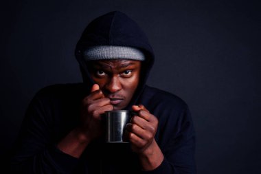 Üzgün görün, elinde metal turist termosu tutan Afro-Amerikan adam kötü hissediyor. Gri şapka ve siyah kapüşonlu stüdyo siyah arka plan giyen turist alkoliği.
