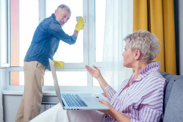 Bella donna bionda seduta sul divano e al lavoro con il computer portatile, il suo uomo in pensione finestra di lavaggio in soggiorno — Foto Stock