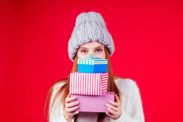 Rothaarige Ingwerfrau trägt stylische Wollmütze und weißen Pullover, blickt in die Kamera und versteckt sich hinter drei Schachteln mit Geschenken in rotem Hintergrund — Stockfoto