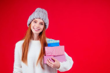 Kızıl saçlı kadın şık yün şapka ve beyaz kazak giyer kameraya bakar ve elinde üç kutu hediye tutar stüdyo kırmızı arka planda kopya yazar