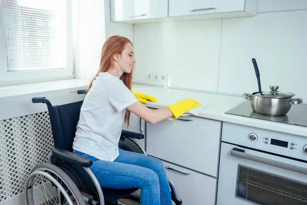 Genç, kızıl saçlı, tekerlekli sandalyede sakat bir kadın. Sarı kurdele eldiveni takıyor ve evi temizliyor. — Stok fotoğraf