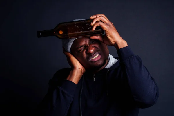 Africano americano homem segurando uma garrafa de vinho e sentindo dor de cabeça merda no estúdio preto background.bum sem-teto afogando a dor — Fotografia de Stock