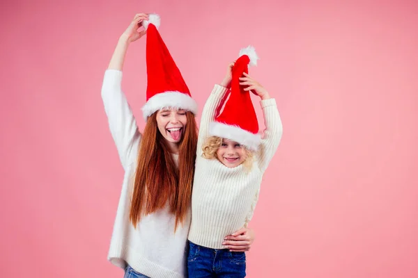 ,Mutlu kızıl saçlı kadın ve şirin sarışın kız Noel Baba şapkasının altında gözleri saklıyor ve stüdyo pembe arka planda birlikte eğleniyorlar. — Stok fotoğraf