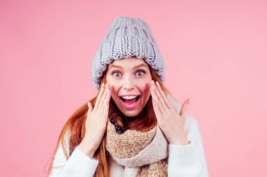 Kızıl saçlı kadın şık yün şapka, kazak ve eşarp giyer kameraya bakar ve yanaklarına dokunur. Stüdyo pembe arka planına krem sürerler.