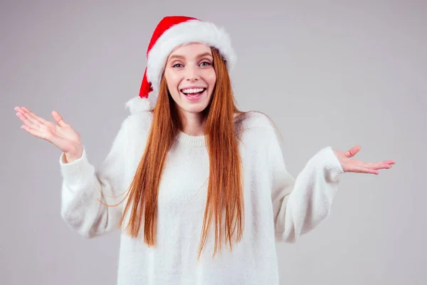 Schöne rothaarige Ingwerfrau mit weißem Strickpullover und Weihnachtsmann-Hut, die Geste im Studio-Backhround zeigt — Stockfoto