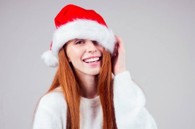 Utangaç, kızıl saçlı, beyaz örgü süveter giyen ve Noel Baba şapkalı stüdyo arka planında saklanan güzel bir kadın.