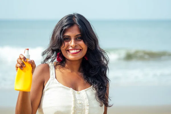 Портрет красивой и улыбающейся белоснежной улыбки индианки с черными вьющимися волосами и темной кожей в белой футболке с бутылкой солнцезащитного спрея на пляже. — стоковое фото