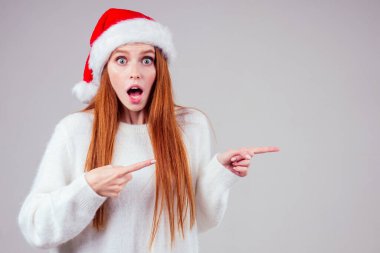 Güzel kızıl saçlı, beyaz örgü süveter giyen ve stüdyo arka planında Noel Baba şapkası takan kadın fotokopi spazmını işaret ediyor.