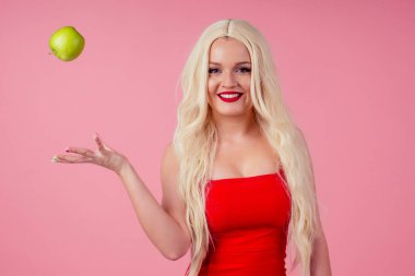 Pembe stüdyo arka planında yeşil elmalı gülümseyen sarışın kadının portresi.