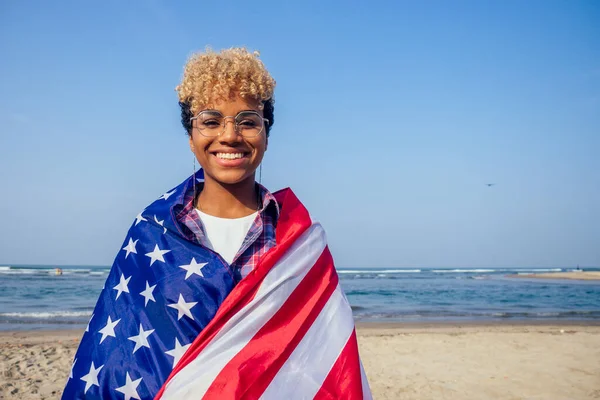 Heureuse femme afro américaine tenant le drapeau des Etats-Unis à la plage tropicale ensoleillée.4 juillet Independence Day concept — Photo