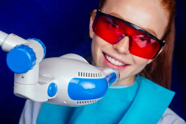 Pelirroja pelirroja joven alegre cliente femenino con gafas rojas de protección sentado en el consultorio dental y conseguir blanqueamiento sonrisa en la silla de huevo — Foto de Stock