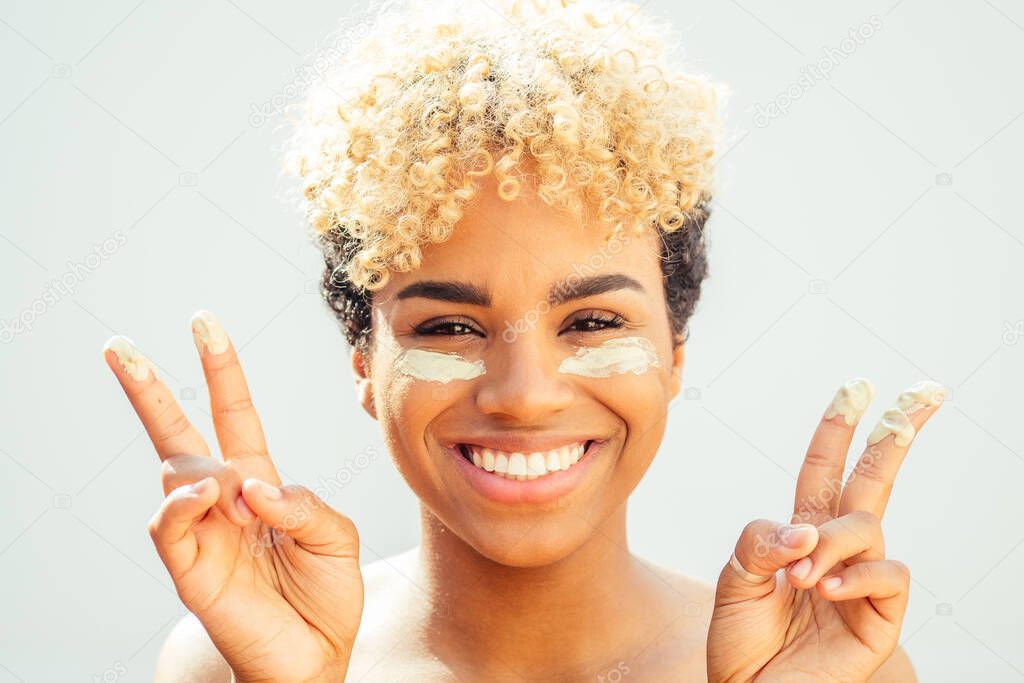 Happy latin brazilian woman with healthy skin applying tone cream on her face on white studio background. she covers bags under the eyes