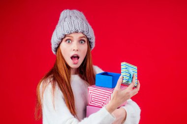 Kızıl saçlı kadın şık yün şapka ve beyaz kazak giyer kameraya bakar ve elinde üç kutu hediye tutar stüdyo kırmızı arka planda kopya yazar