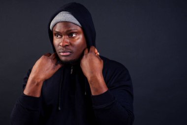 Yakışıklı Afro-Amerikalı erkek gri şapkalı ve kapüşonlu siyah kapüşonlu kamera stüdyosunun arka planına ciddi ve güçlü bir bakış atıyor.
