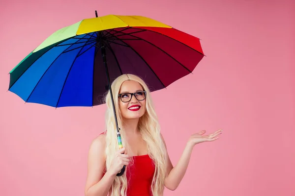 Όμορφη χαμογελαστή νεαρή γυναίκα με μακριά ξανθιά περούκα κρατώντας πολύχρωμη ομπρέλα στο στούντιο ροζ φόντο — Φωτογραφία Αρχείου