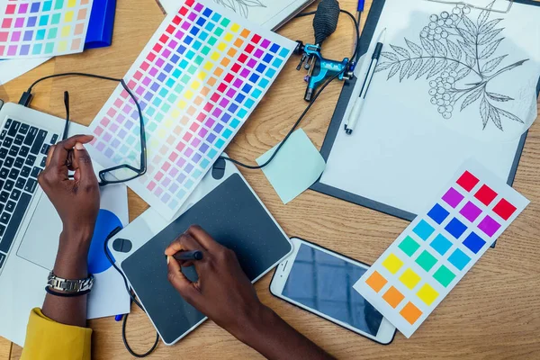Hands of afro american woman in a stylish yellow jacket and multi-colored dreadlocks pigtails is graphic designer draws a sketch on a tablet in workplace in a light large modern office — Stock Photo, Image