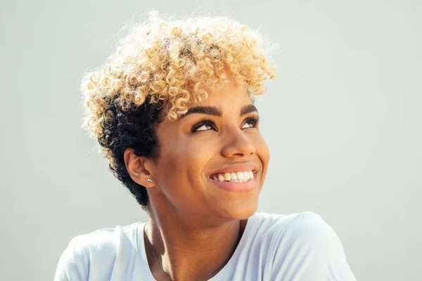 Hispanic latin woman with afro blonde haircut dressed in white top looking up with charming shy smile in studio white background — Stock Photo, Image