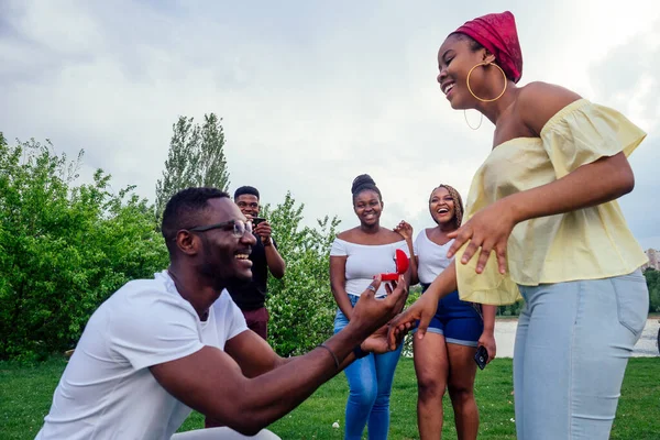 American male standing on one knee asking woman marriage giving ring proposing surprised shocked while gathering group friends all around in shock confusing evening cloudy autumn spring in the park — Stock Photo, Image