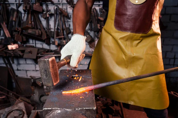 Kalapácsipar kis üzleti koncepció.afro-amerikai férfi öltözött történelmi ruhák kalapácsol az üllő. Egy kovács fémterméket kovácsol. — Stock Fotó