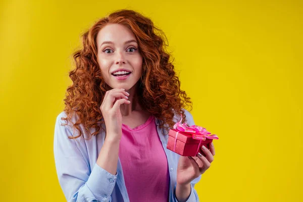 Rödhårig lockigt ingefära kvinna som håller presentförpackning i studio gul bakgrund. mödrar dag och åttonde marsor St. Alla hjärtans dag koncept — Stockfoto